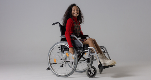 Menina sorridente sentada em cadeira de rodas recebida por meio de doação de equipamentos hospitalares, em ambiente iluminado, representando acolhimento e inclusão.