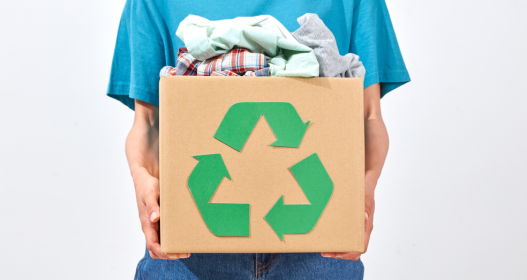 Pessoa segurando caixa de papelão com símbolo de reciclagem e roupas, representando doação de roupas e consumo consciente.