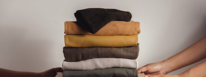 Pilha de roupas dobradas sendo entregue por mãos solidárias, simbolizando doação de roupas com cuidado e respeito.